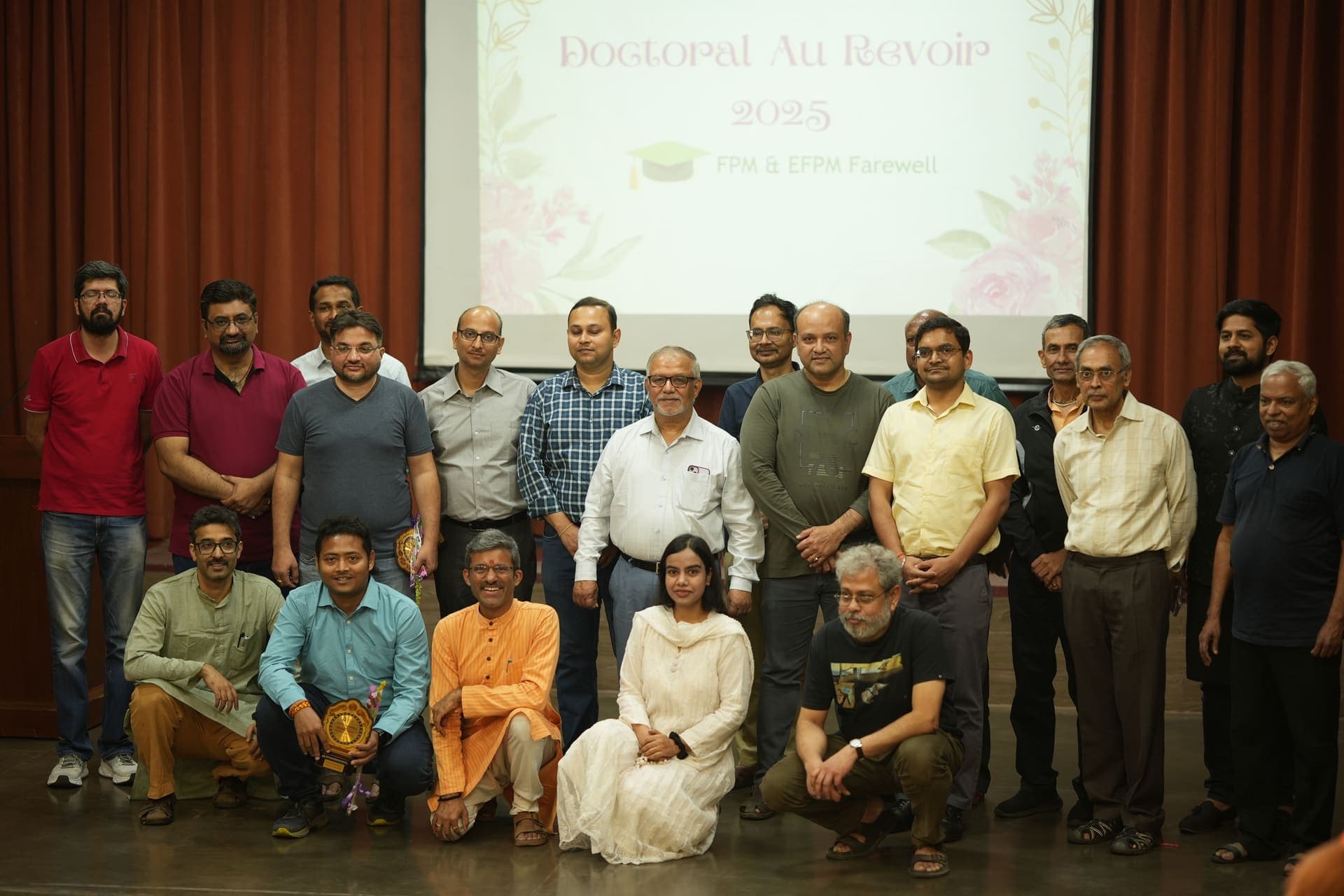 A group photo of FPM and EFPM scholars and faculty on stage at XLRI's Doctoral Au Revoir 2025, with the event title on screen behind them
