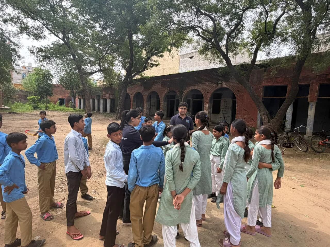 Management students listening to schoolchildren in a shaded village courtyard